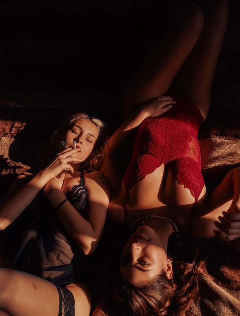 Two women in lingerie lying on a brown surface, one smoking. High-angle shot, dark lighting creates shadows. Lingerie fashion photography.