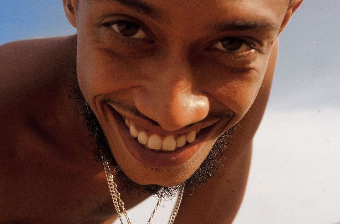 Portrait of a dark-skinned adult male with a beard stubble smiling broadly at the camera, featuring white teeth, clear skin, and a gold chain.
