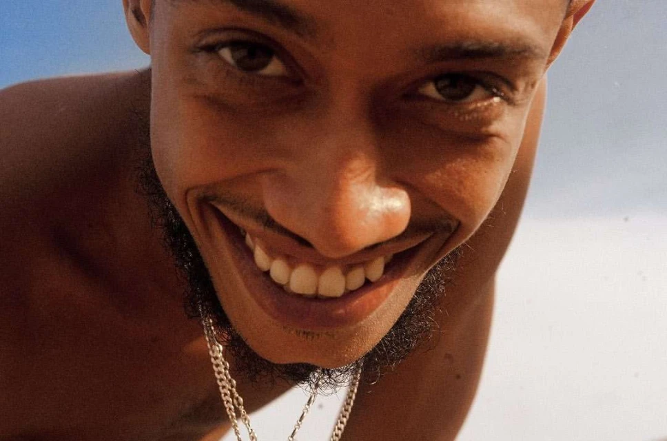 Portrait of a dark-skinned adult male with a beard stubble smiling broadly at the camera, featuring white teeth, clear skin, and a gold chain.