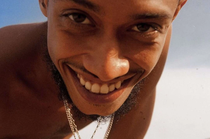 Portrait of a dark-skinned adult male with a beard stubble smiling broadly at the camera, featuring white teeth, clear skin, and a gold chain.