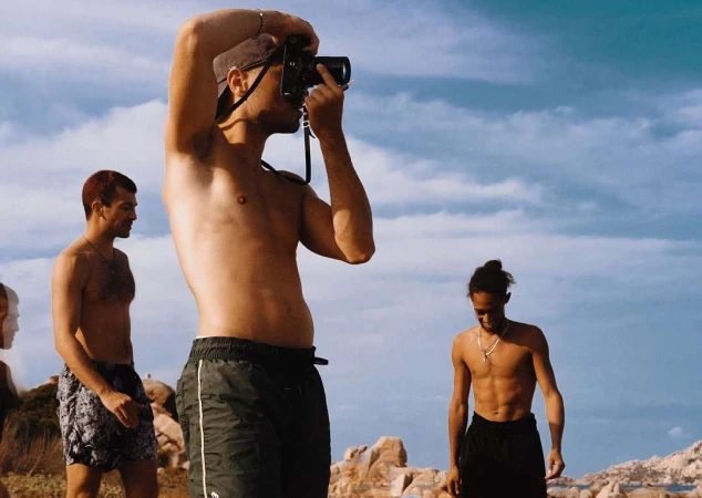 Three young men shirtless on a sandy beach, one holding a vintage camera to his eye against a sunny blue sky