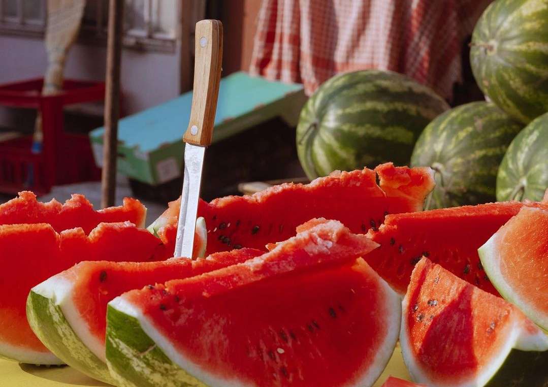 Close-up of sliced watermelon with knife in a market stall, featuring red flesh, green rind, and scattered seeds. Summer fruit still life.