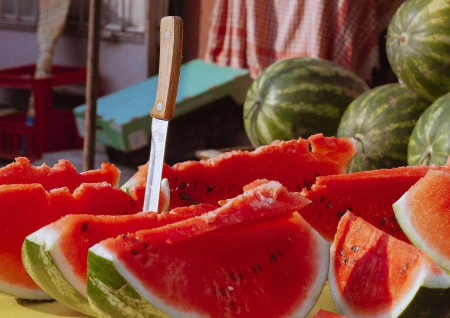 Close-up of sliced watermelon with knife in a market stall, featuring red flesh, green rind, and scattered seeds. Summer fruit still life.