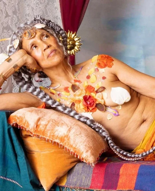 Older woman with braids and flowers poses in a recumbent position with head resting on hand, draped across striped bedding.