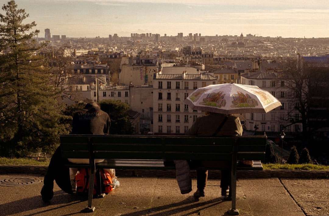 Two people on a park bench overlooking a cityscape with an umbrella. Daytime view of the buildings in an urban environment.