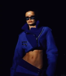 Model in dark sunglasses wearing a vibrant blue cropped jacket against a dark background, photographed by Anais Stupka.