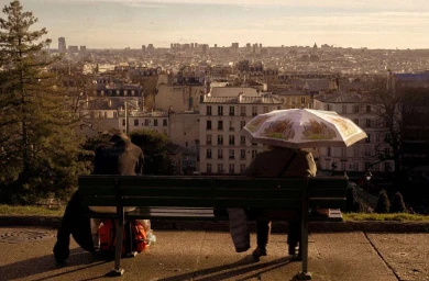 Two people sitting on a green park bench overlooking a distant cityscape under warm sunlight, photographed by Anais Stupka.