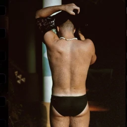 Rear view of a man wearing dark swim briefs and a pearl necklace while removing a black shirt, photographed in London.
