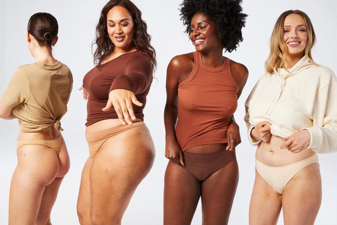 Four diverse women wearing neutral-tone underwear and tops stand against a white background, smiling, showcasing body positivity and fashion.