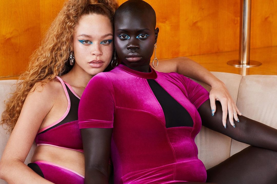 Two women pose indoors on a cream couch; one with curly red hair and one with a shaved head, wearing matching pink and black outfits.