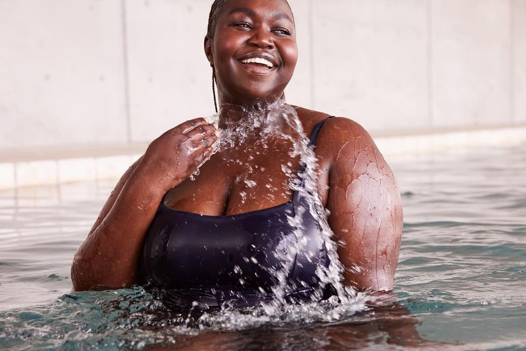 Smiling African American woman in a dark swimsuit stands in a swimming pool, splashing water with her hand under bright lighting.