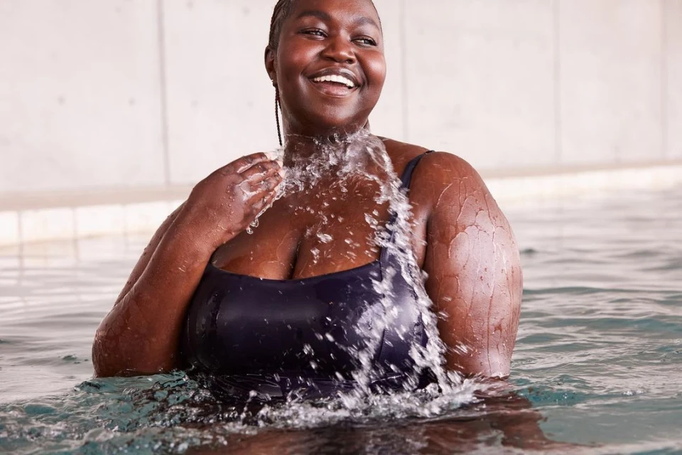 Smiling African American woman in a dark swimsuit stands in a swimming pool, splashing water with her hand under bright lighting.