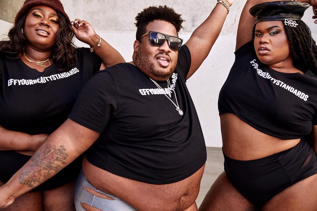 Three plus-size models wearing black T-shirts with the text "EFFYOURBEAUTYSTANDARDS" pose in front of a light-colored concrete wall.