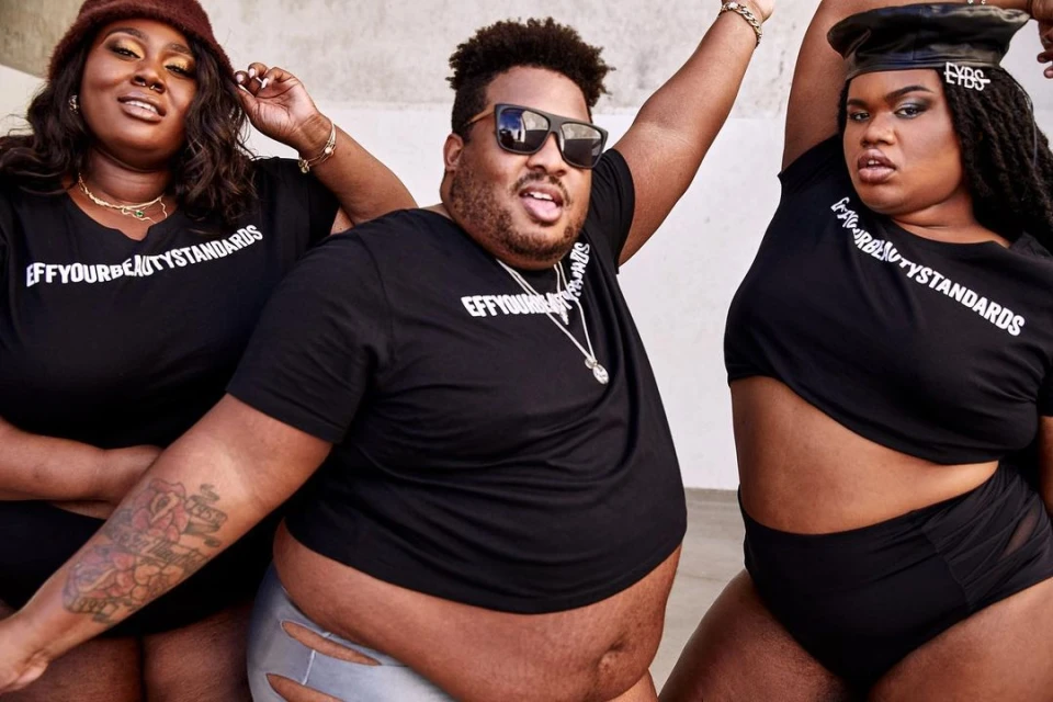 Three plus-size models wearing black T-shirts with the text "EFFYOURBEAUTYSTANDARDS" pose in front of a light-colored concrete wall.
