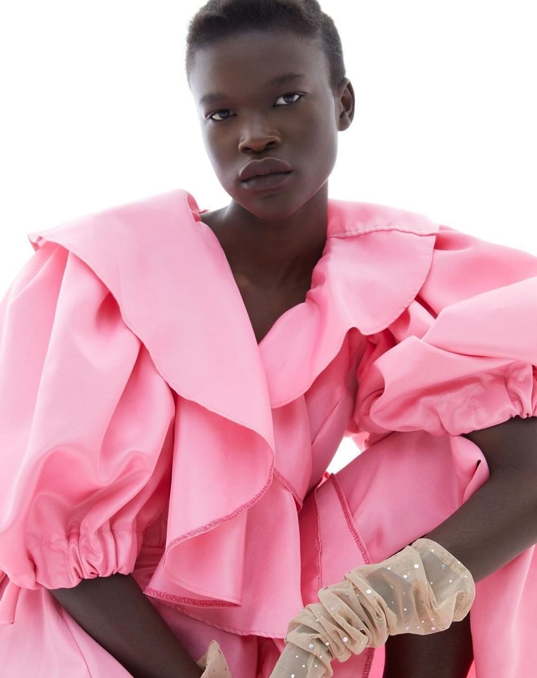 Adult female with dark skin poses in a studio, wearing a large ruffled pink dress and sheer, embellished arm coverings in portrait shot.