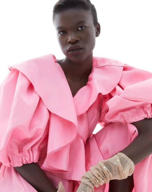 Adult female with dark skin poses in a studio, wearing a large ruffled pink dress and sheer, embellished arm coverings in portrait shot.