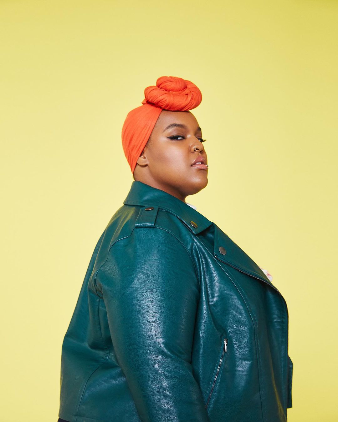 Portrait of an adult female with an orange head wrap and teal leather jacket against a yellow background in a photo studio.