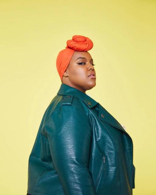 Portrait of an adult female with an orange head wrap and teal leather jacket against a yellow background in a photo studio.