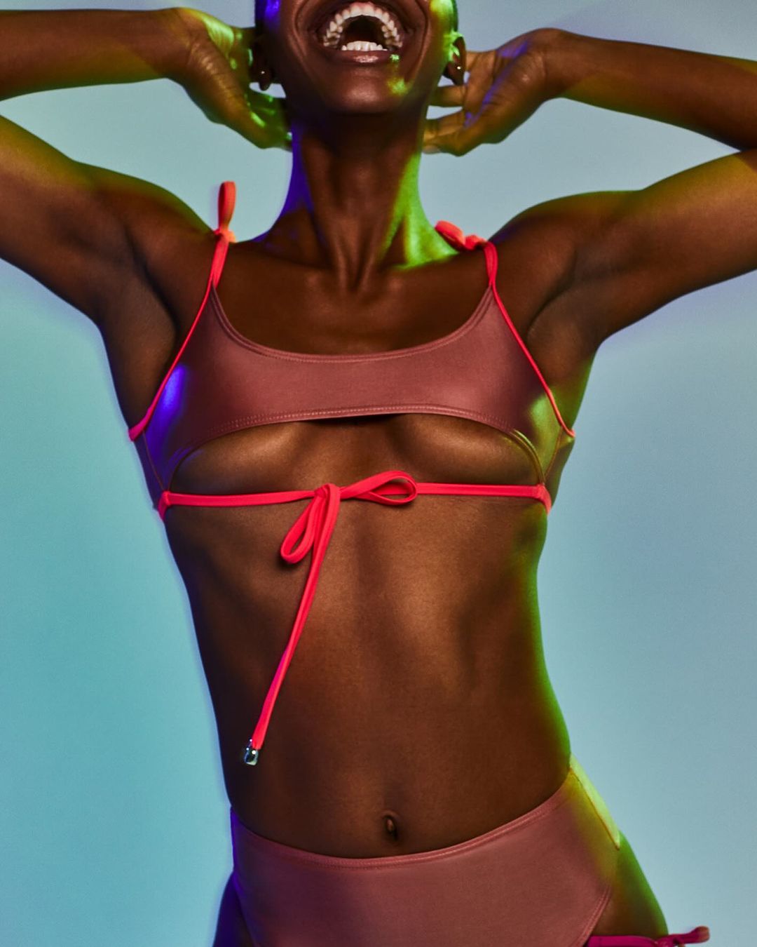 A dark-skinned female model smiles broadly, wearing a two-piece rose-colored swimsuit with coral straps against a vibrant blue and green background.