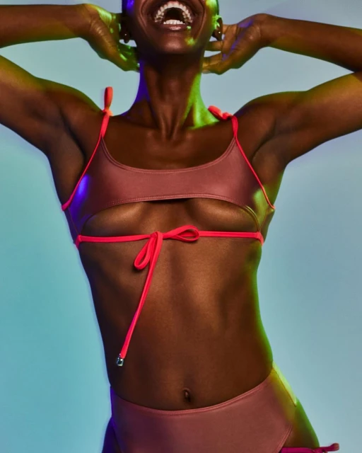 A dark-skinned female model smiles broadly, wearing a two-piece rose-colored swimsuit with coral straps against a vibrant blue and green background.
