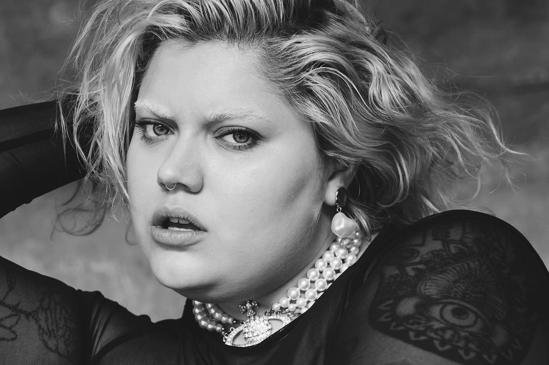 Black-and-white portrait of a fair-skinned woman with bleached hair, wearing a sheer top and pearl jewelry in a studio setting.