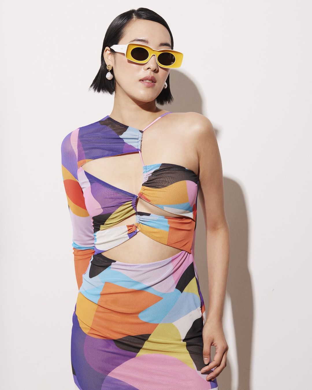 Young woman with short dark hair wears a colorful cutout dress and retro yellow sunglasses in a fashion studio shot against white.