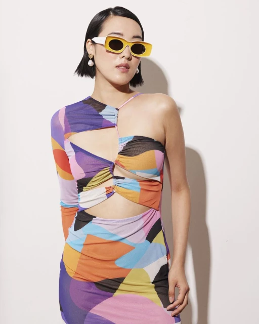 Young woman with short dark hair wears a colorful cutout dress and retro yellow sunglasses in a fashion studio shot against white.