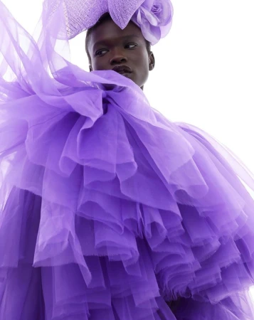 Model in voluminous purple tulle ruffled dress with matching headpiece against a white background