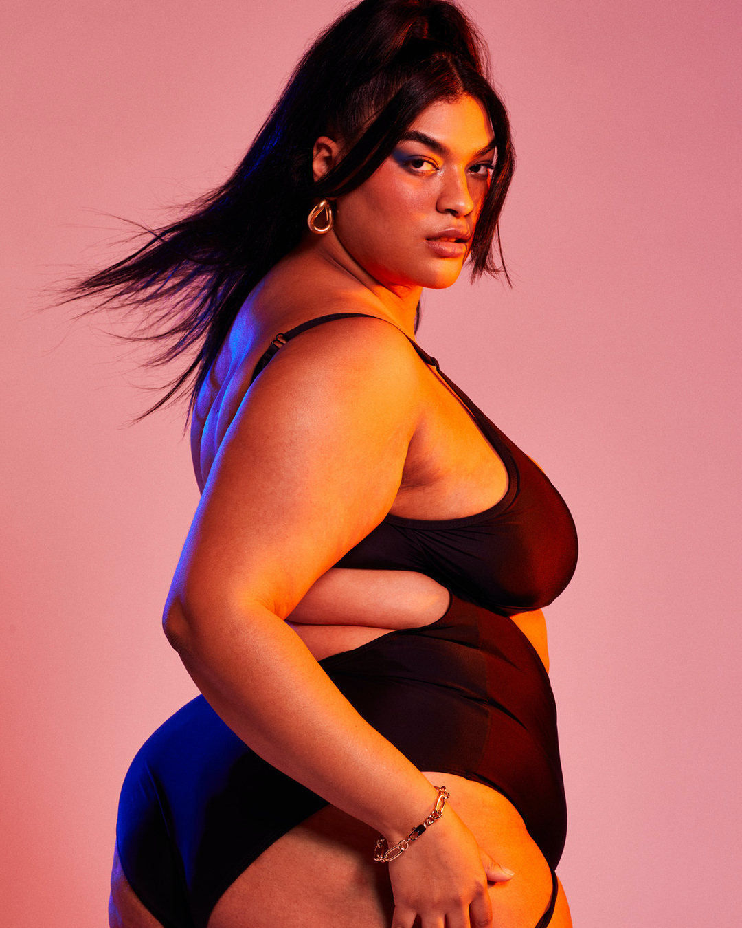 Portrait of a plus-size adult woman wearing a black bra and underwear set, posing in a studio with dynamic orange and pink lighting.