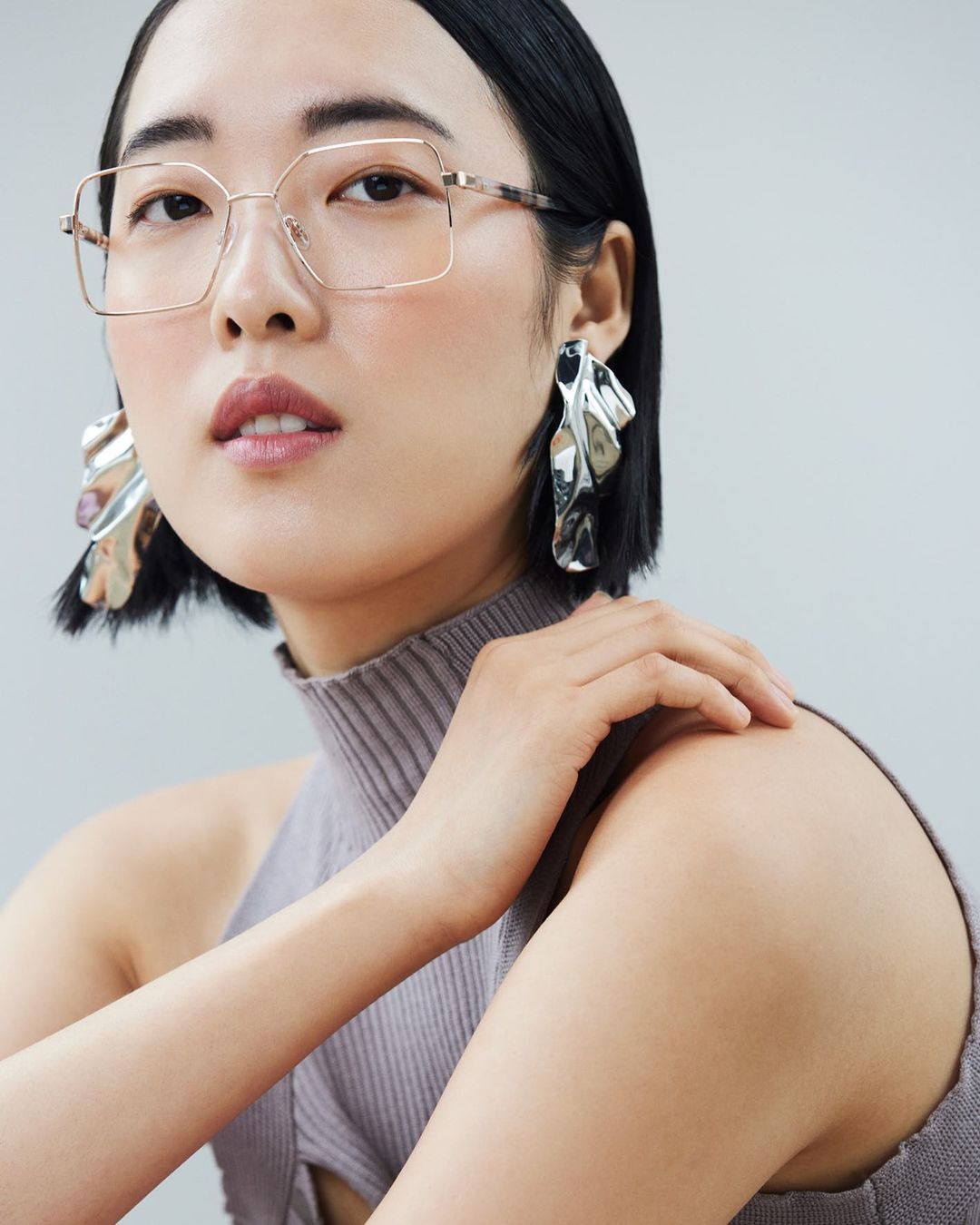 Portrait of a stylish Asian woman wearing glasses and large statement earrings against a white background in a studio setting.
