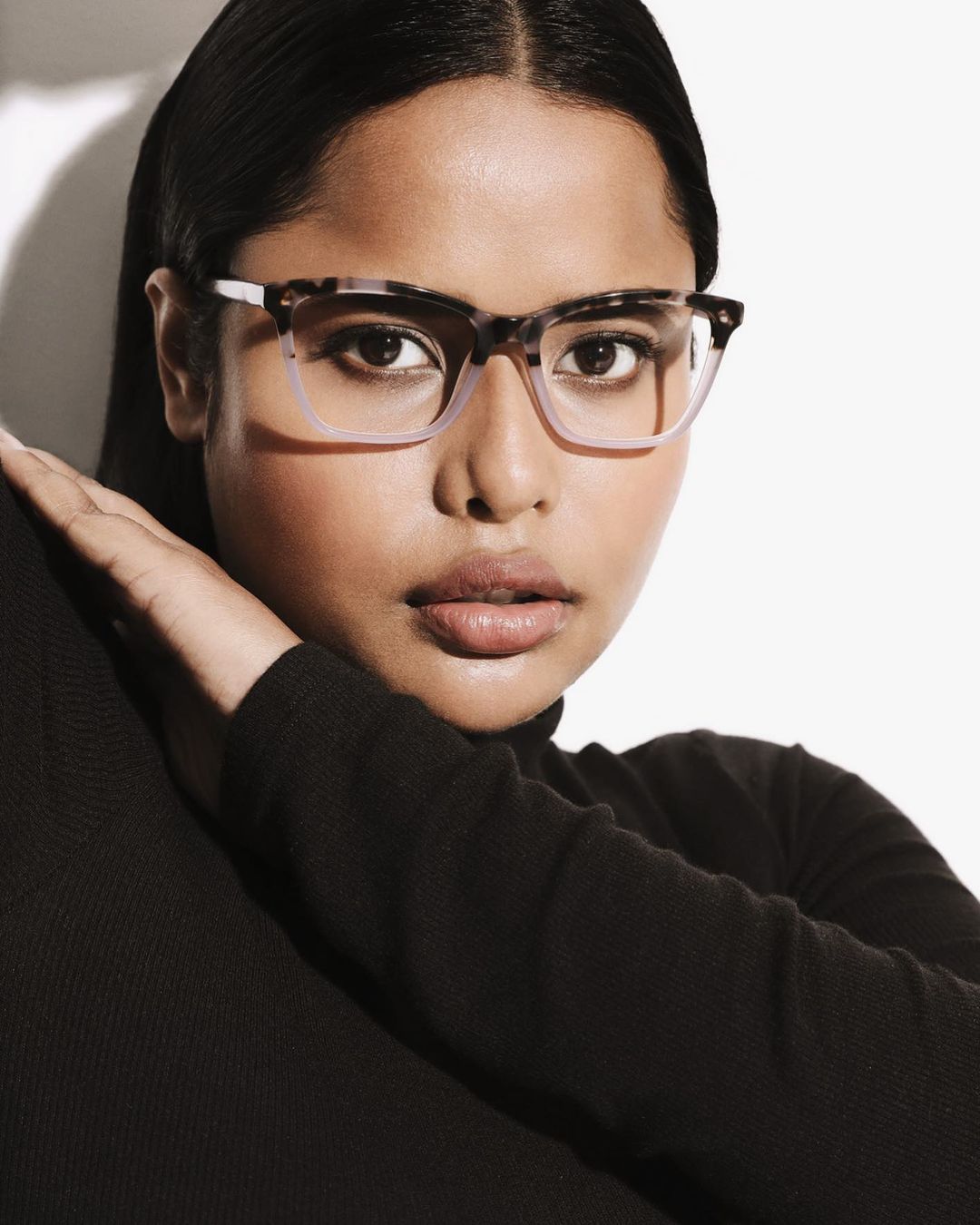 Close-up portrait of a beautiful adult woman with tan skin wearing black turtleneck and eyeglasses against a bright white backdrop.