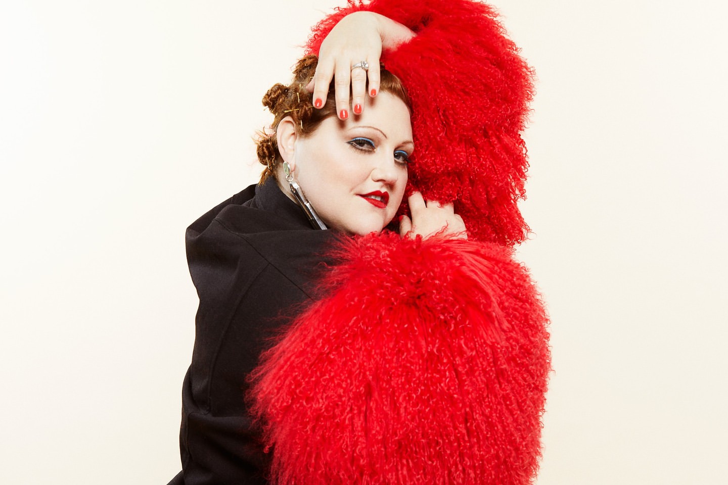 Portrait of a stylish woman with red curly hair and a large fuzzy red collar, wearing a black coat, in a photo studio setting.
