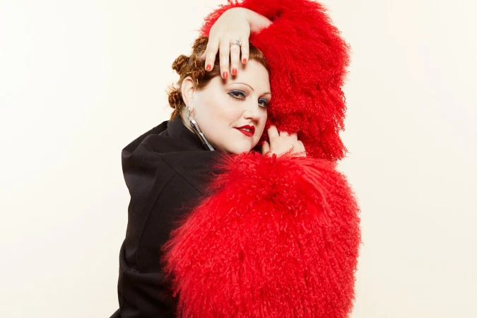 Portrait of a stylish woman with red curly hair and a large fuzzy red collar, wearing a black coat, in a photo studio setting.
