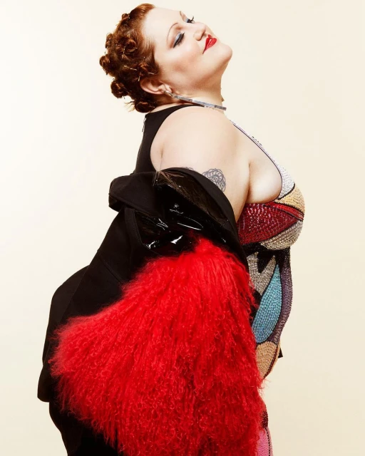 Portrait of a curvy woman with auburn hair and red lipstick wearing a colorful beaded top with a black jacket and red faux fur stole.