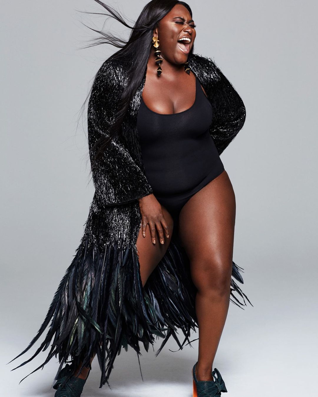 Full-length portrait of a Black woman in a black bodysuit and feather robe against a grey background. She is laughing with an open mouth.