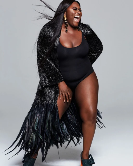 Full-length portrait of a Black woman in a black bodysuit and feather robe against a grey background. She is laughing with an open mouth.
