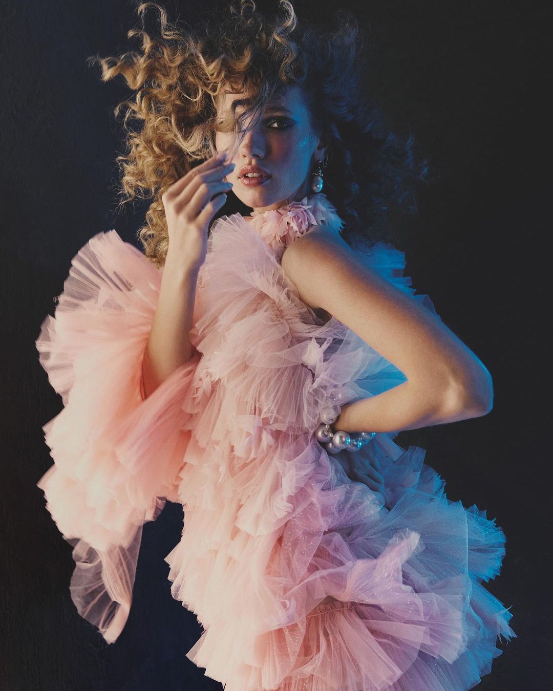 Woman in pink ruffled dress poses against a dark background with blue lighting accents, showcasing fashion and style.