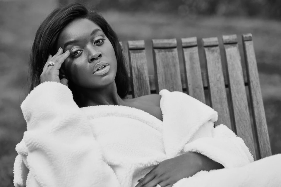 Black and white portrait of a woman in a white robe, lying on a wooden chair, gazing at the camera with a serene expression.