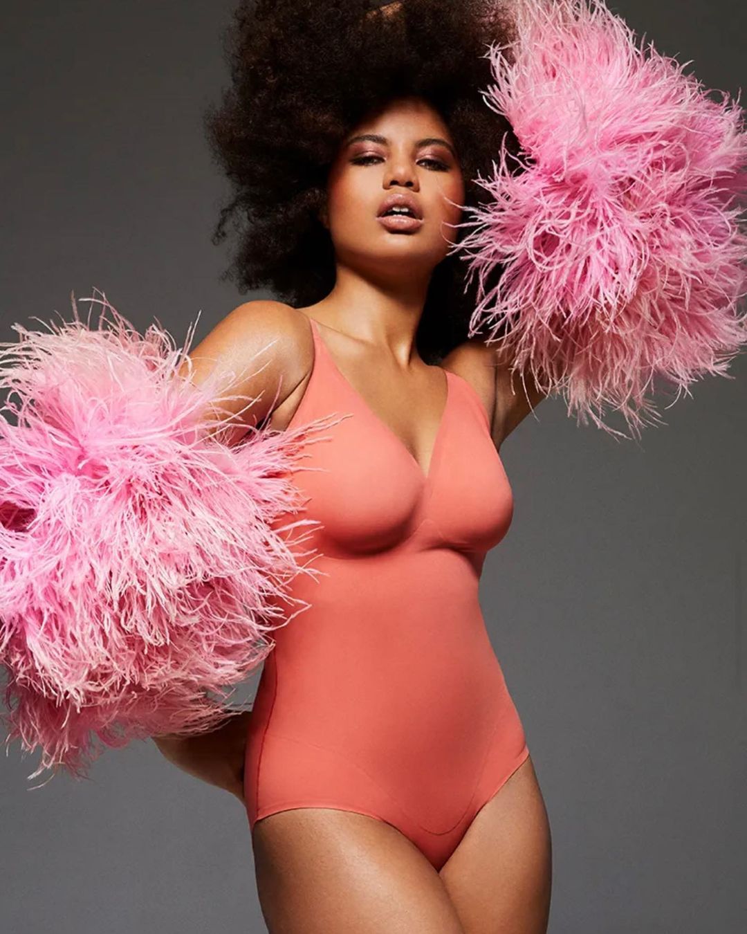 Portrait of an adult female posing in a coral leotard with large, fluffy pink feather boas against a grey backdrop in a studio setting.