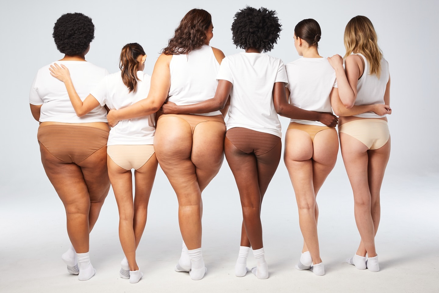 Diverse group of women in underwear and t-shirts stand with their arms around each other in a studio, showcasing body positivity.