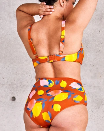 A woman from behind in a patterned two-piece swimsuit featuring a vibrant design with lemons and blue leaves against a terracotta background.