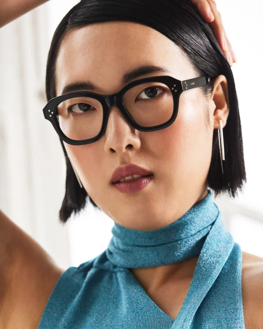 Close-up of a woman with black framed glasses and a shiny blue top. She has black hair and her hand is near her face.