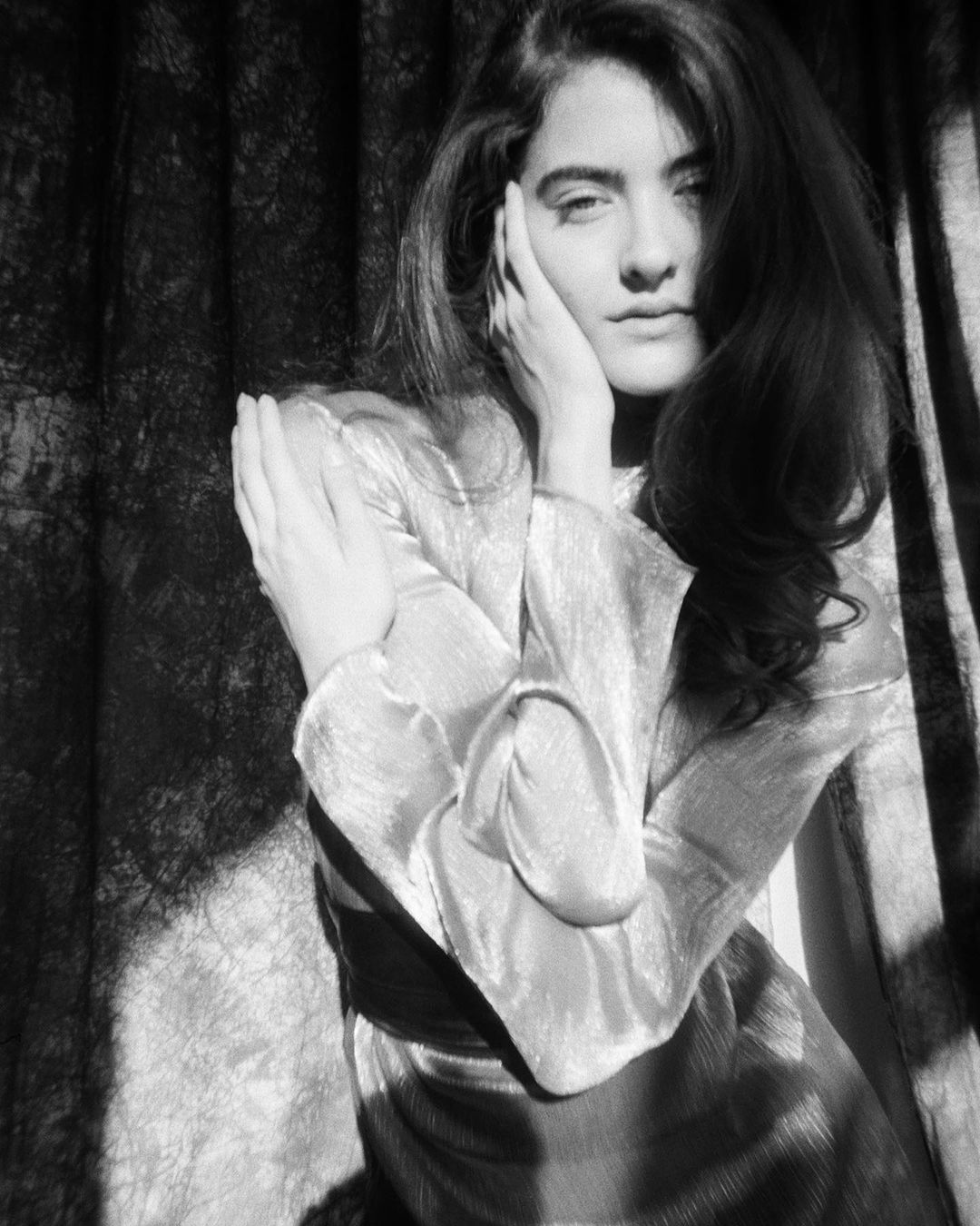 Monochrome portrait of a woman with long hair in a satin dress, posing with hands near her face against a draped textile backdrop.