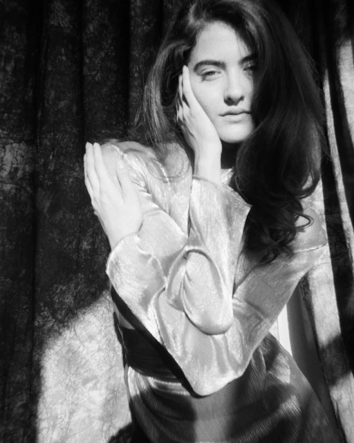 Monochrome portrait of a woman with long hair in a satin dress, posing with hands near her face against a draped textile backdrop.