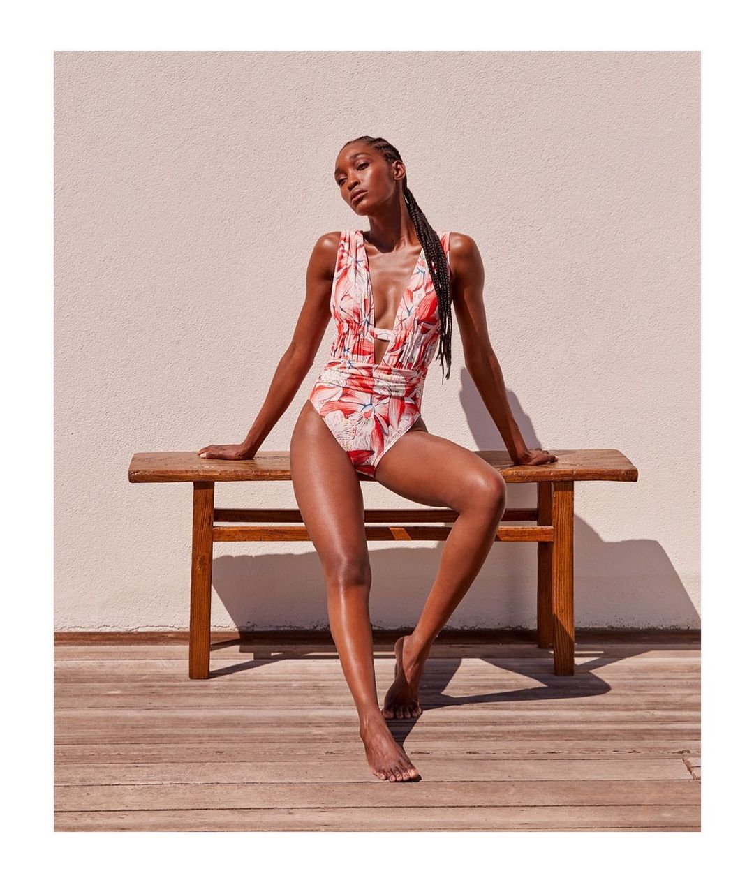 Black woman wearing a red floral one-piece swimsuit seated on a wooden bench against a light wall by Anastasia Garcia