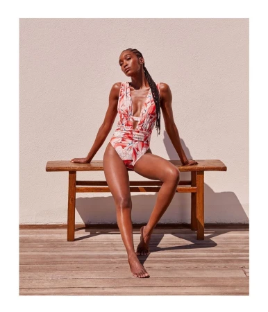 Black woman wearing a red floral one-piece swimsuit seated on a wooden bench against a light wall by Anastasia Garcia