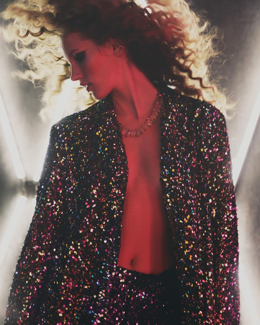 Woman in sequin blazer with curly hair poses in front of bright geometric lights. Fashion editorial style, beauty shot, indoor setting.