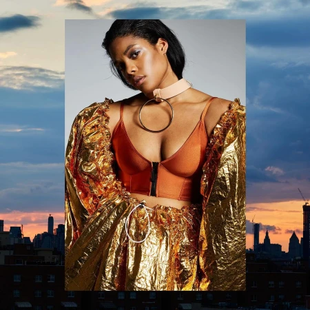 Fashionable adult woman with gold skirt and orange top poses in front of urban skyline at dusk with sunset, wearing matching choker.