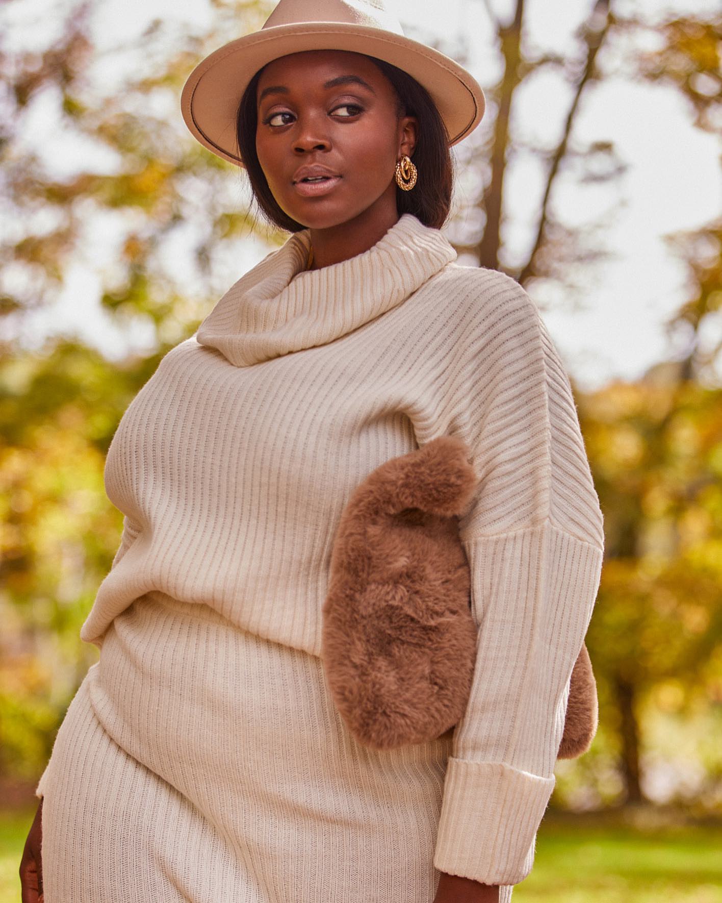 A single adult woman wearing a cream-colored sweater dress and hat stands in an outdoor environment with autumn foliage in the background.