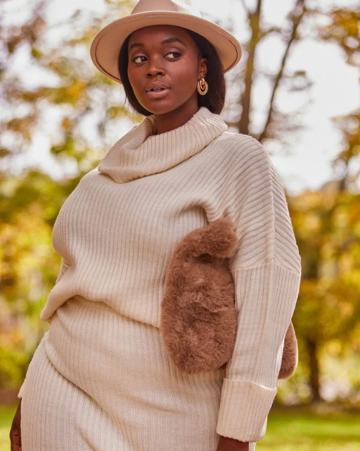 A single adult woman wearing a cream-colored sweater dress and hat stands in an outdoor environment with autumn foliage in the background.